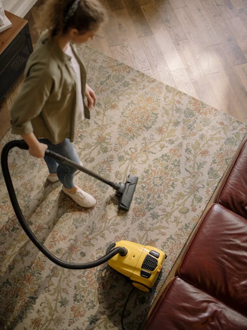A yellow vacuum cleaner with a long black hose and a floor attachment is positioned on a patterned area rug in a living room, adjacent to a wooden sideboard with black metal legs. The sideboard holds 