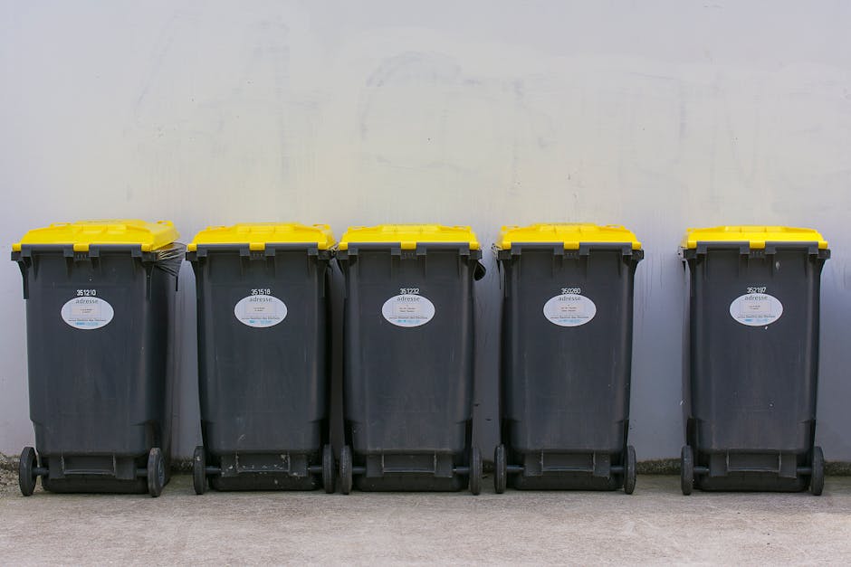 A person dressed in orange protective overalls is standing on a grey industrial-style floor, holding two large blue plastic garbage bags filled with waste. The silhouette of the person's legs and whit