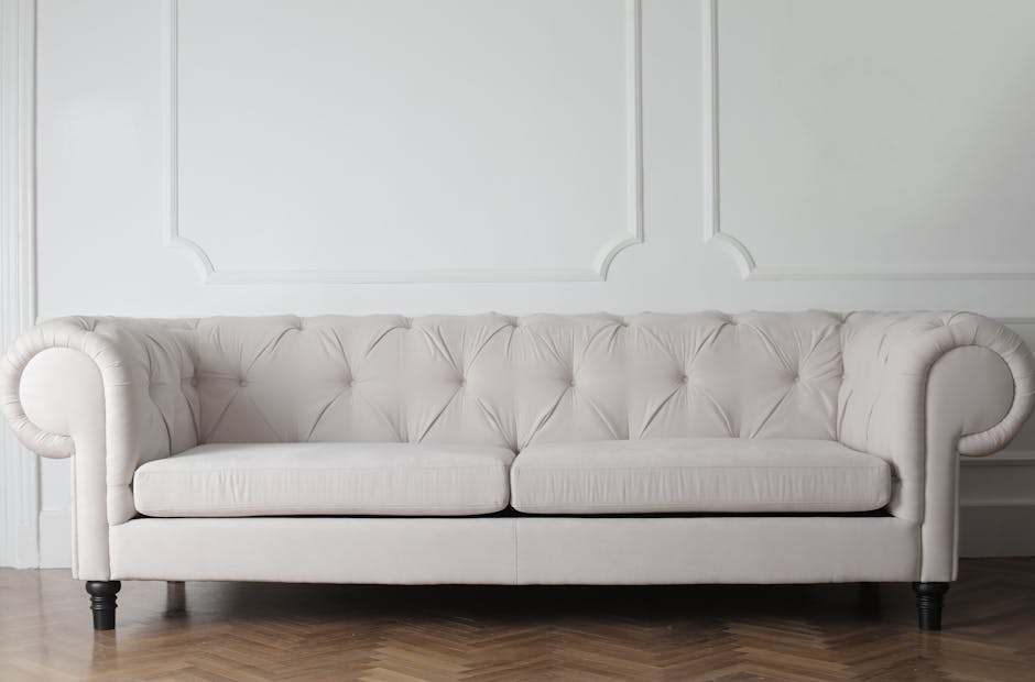 A person wearing black gloves is using a handheld electric upholstery cleaner on a dark grey fabric sofa located in a living room. The sofa has a modern design with wide armrests and is positioned on
