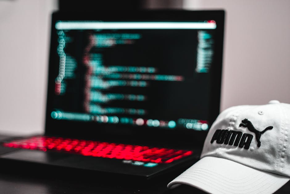 Close-up of a silver MacBook laptop on a white desk displaying lines of coding on its dark screen, with a white smartphone placed in front of it and an open notebook with a silver pen resting on it to
