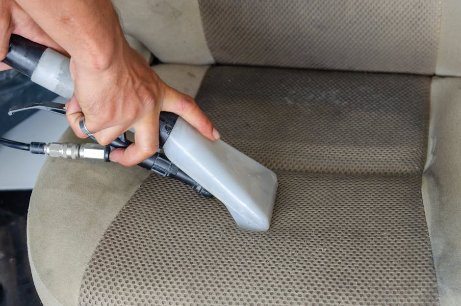Close-up of a hand holding a handheld steam cleaner or upholstery cleaning device, using it to clean a beige fabric car seat with a textured, patterned surface. The nozzle is pressed against the fabri